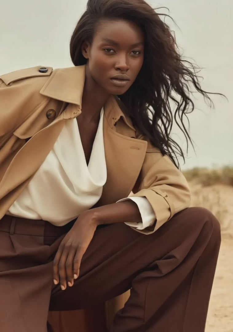 Model wearing a camel trench coat and brown oversized trousers crouches against a desert background, looking intently at the camera.