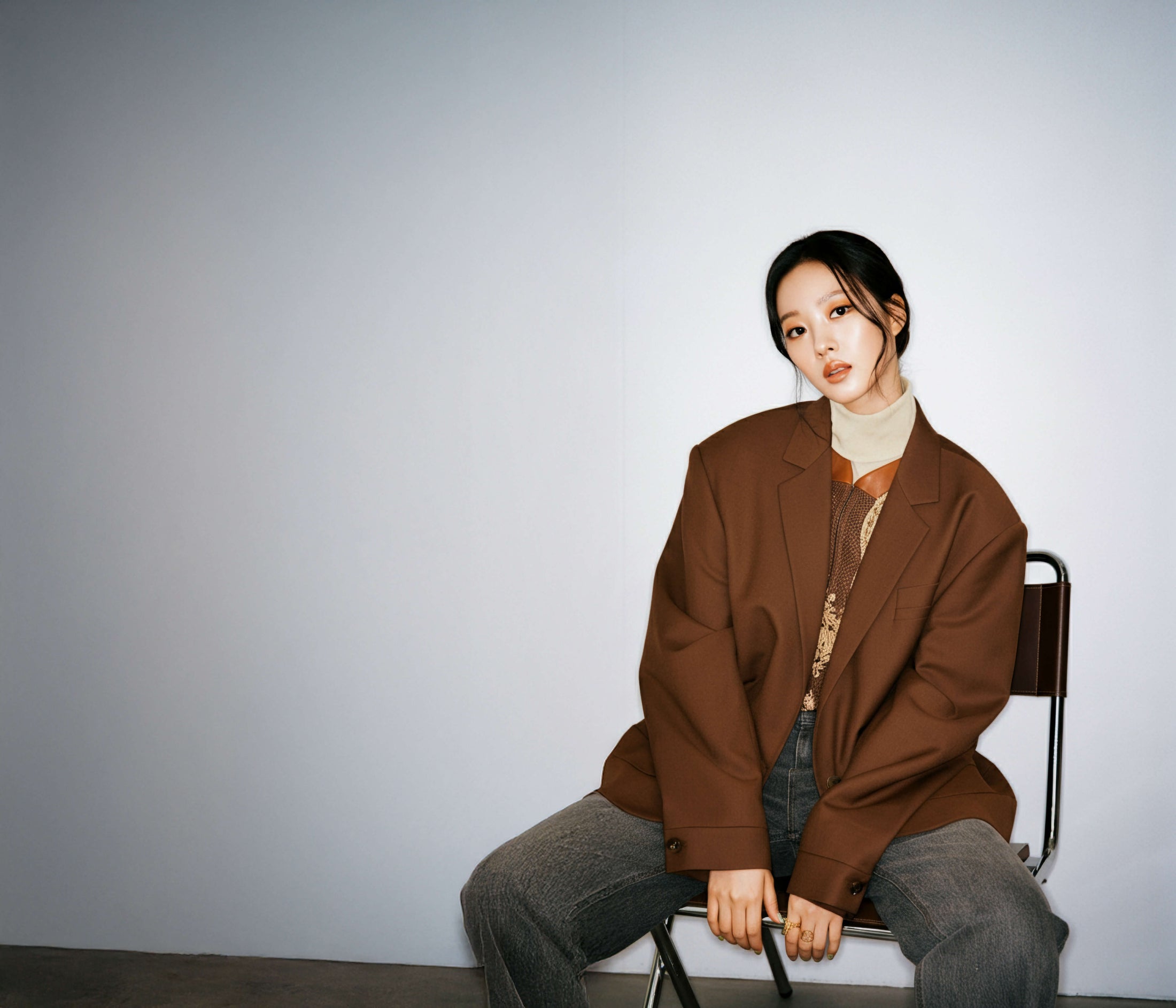 Woman sits against bare background wearing oversized brown blazer and gray jeans.