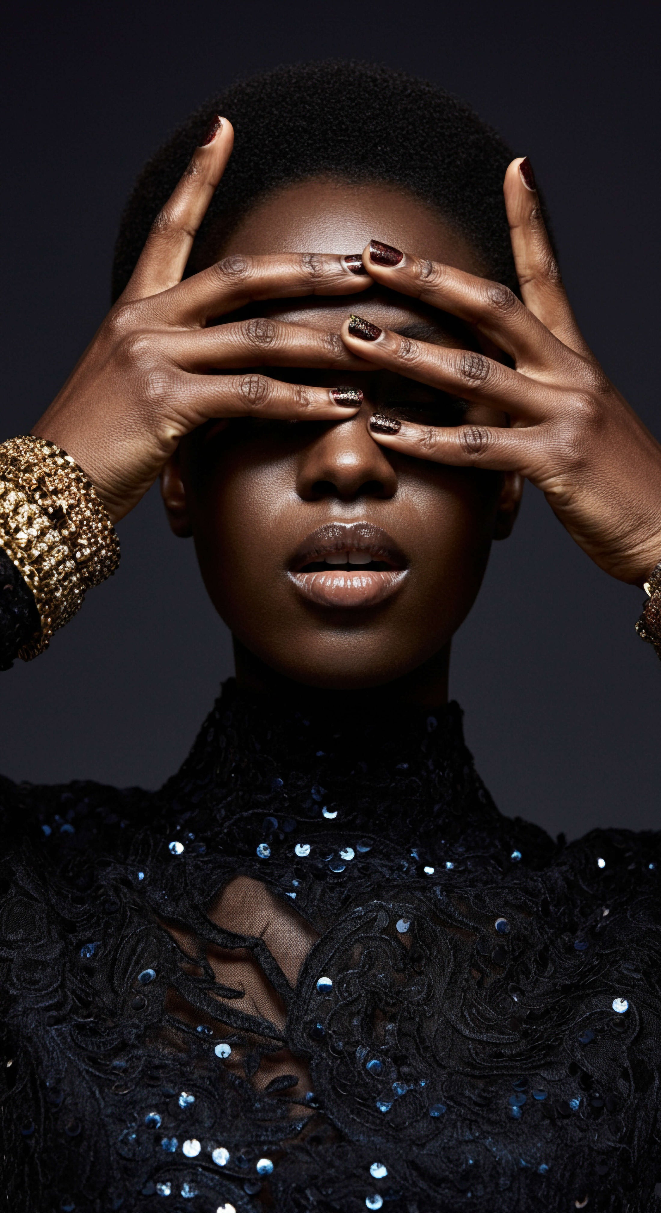 Model wearing black dress with sequins and gold bangles covers her eyes.